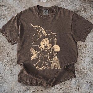 Puede incluir: Camiseta marr&oacute;n con un dise&ntilde;o beige de Minnie Mouse vestida de bruja, sosteniendo una escoba. El dise&ntilde;o tiene un aspecto vintage y desgastado. La camiseta tiene mangas cortas y cuello redondo, ideal para uso diario.