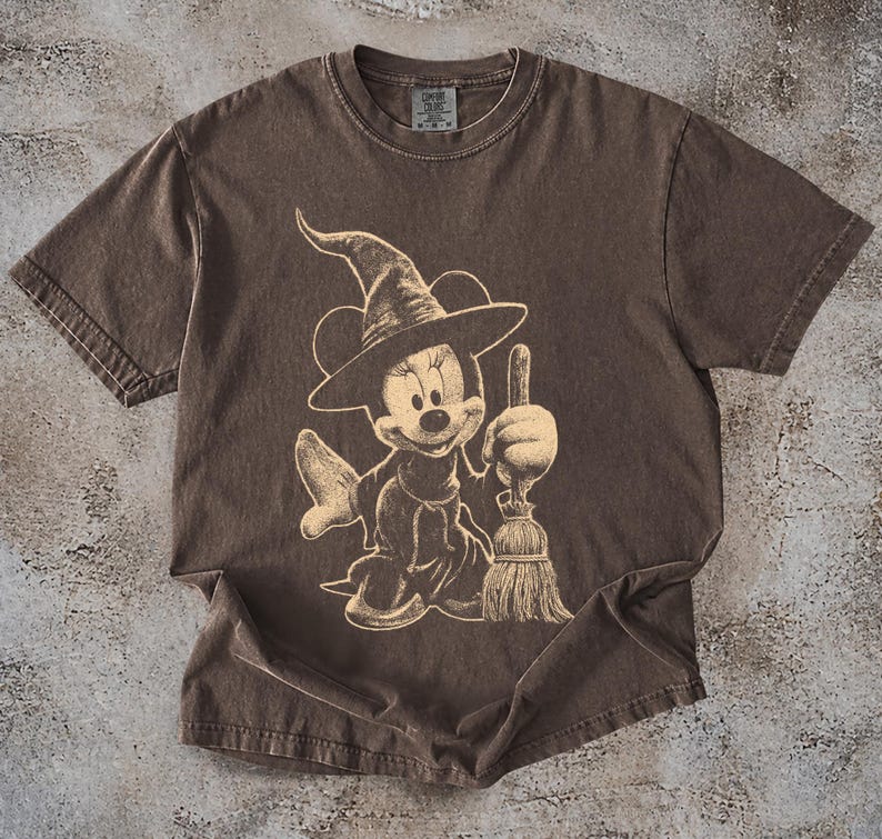 Puede incluir: Camiseta marr&oacute;n con un dise&ntilde;o beige de Minnie Mouse vestida de bruja, sosteniendo una escoba. El dise&ntilde;o tiene un aspecto vintage y desgastado. La camiseta tiene mangas cortas y cuello redondo, ideal para uso diario.