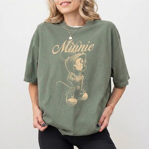 May include: An olive green short-sleeve t-shirt featuring a beige graphic print of the character Minnie Mouse wearing a hoodie and sneakers. The word Minnie is printed in a cursive script above the character illustration on the front of the shirt.