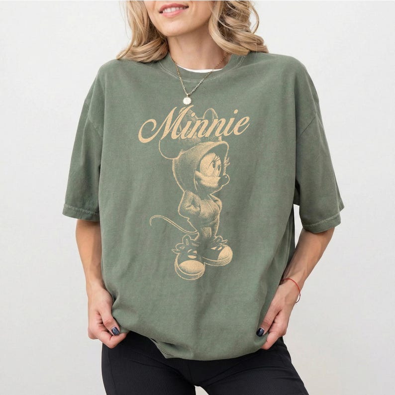 May include: An olive green short-sleeve t-shirt featuring a beige graphic print of the character Minnie Mouse wearing a hoodie and sneakers. The word Minnie is printed in a cursive script above the character illustration on the front of the shirt.