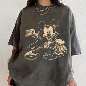 May include: Dark gray t-shirt featuring a vintage-style illustration of Mickey Mouse dressed as a vampire, holding a jack-o'-lantern. The design is in shades of beige and black. The shirt has short sleeves and a relaxed fit, perfect for Halloween.