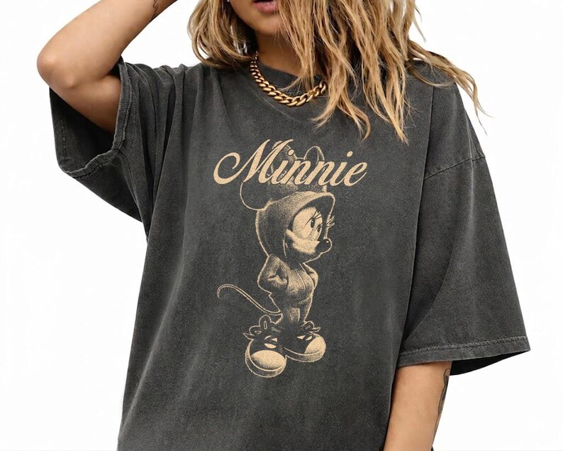 May include: A charcoal gray short-sleeved t-shirt featuring a beige graphic print of Minnie Mouse wearing a hooded sweatshirt and sneakers. The text Minnie is printed in a cursive script above the character illustration on the front of the shirt.