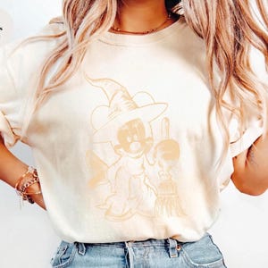 Puede incluir: Camiseta de color marfil con un personaje de dibujos animados disfrazado de bruja, sosteniendo una escoba. El personaje lleva un sombrero de bruja y una capa. El texto "Colors Ivory" est&aacute; en la esquina superior izquierda. La camiseta es de estilo casual.