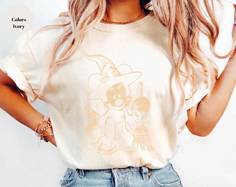 Puede incluir: Camiseta de color marfil con un personaje de dibujos animados disfrazado de bruja, sosteniendo una escoba. El personaje lleva un sombrero de bruja y una capa. El texto "Colors Ivory" est&aacute; en la esquina superior izquierda. La camiseta es de estilo casual.