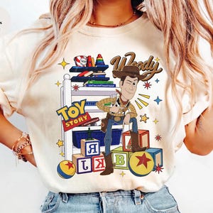 K&ouml;nnte beinhalten: Elfenbeinfarbenes T-Shirt mit einer Cartoon-Illustration von Woody aus Toy Story. Das Design enth&auml;lt die Schriftz&uuml;ge "Woody" und "Toy Story" sowie bunte Bl&ouml;cke, B&uuml;cher und Sterne. Ein l&auml;ssiges Kleidungsst&uuml;ck.