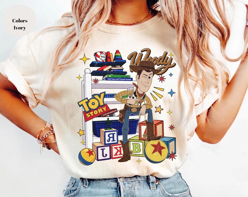 K&ouml;nnte beinhalten: Elfenbeinfarbenes T-Shirt mit einer Cartoon-Illustration von Woody aus Toy Story. Das Design enth&auml;lt die Schriftz&uuml;ge "Woody" und "Toy Story" sowie bunte Bl&ouml;cke, B&uuml;cher und Sterne. Ein l&auml;ssiges Kleidungsst&uuml;ck.