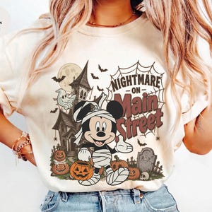 May include: Ivory-colored t-shirt featuring a Halloween-themed graphic. The design includes a cartoon character wrapped in bandages, a haunted house, pumpkins, bats, and the text "Nightmare on Main Street."
