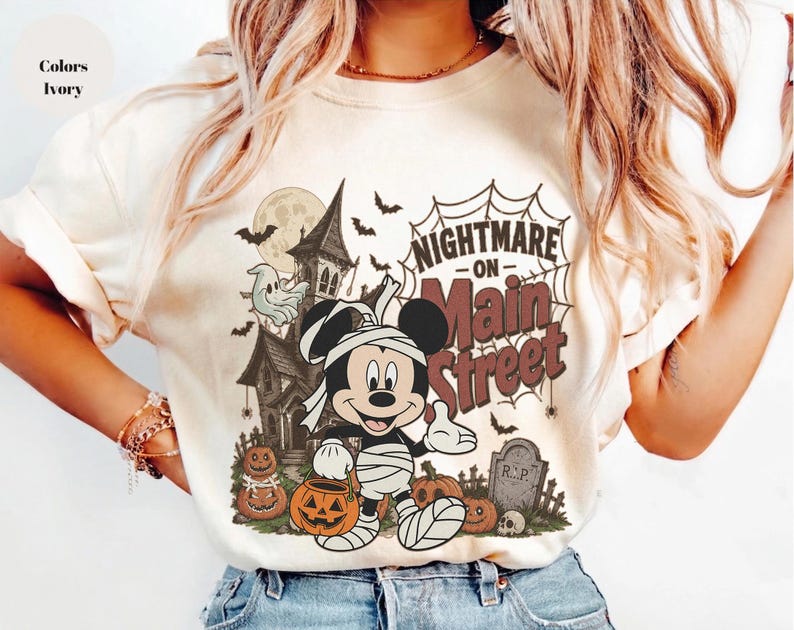 May include: Ivory-colored t-shirt featuring a Halloween-themed graphic. The design includes a cartoon character wrapped in bandages, a haunted house, pumpkins, bats, and the text "Nightmare on Main Street."