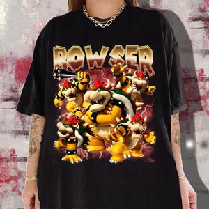 May include: Black t-shirt featuring a graphic of Bowser and several Koopa Troopas with the word "BOWSER" in gold lettering. The design includes red and yellow accents, with lightning bolts in the background. The shirt is a casual wear item.