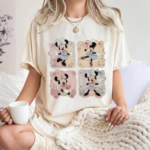 May include: Cream-colored t-shirt featuring a four-panel graphic of Minnie Mouse in various poses. Each panel has a floral background with Minnie in a different outfit. The shirt has short sleeves and a relaxed fit.
