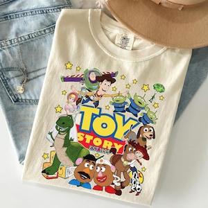 Pu&ograve; includere: T-shirt color crema con una grafica colorata dei personaggi del film d'animazione "Toy Story". Il design include Woody, Buzz Lightyear, Rex e i Little Green Men, con le parole "Toy Story Est. 1995" in giallo e rosso.