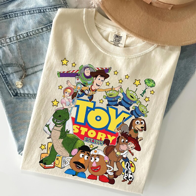 Pu&ograve; includere: T-shirt color crema con una grafica colorata dei personaggi del film d'animazione "Toy Story". Il design include Woody, Buzz Lightyear, Rex e i Little Green Men, con le parole "Toy Story Est. 1995" in giallo e rosso.
