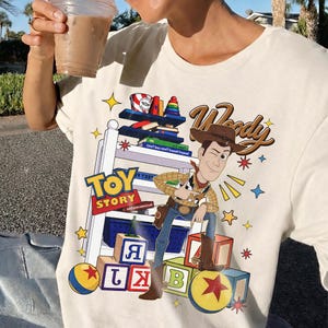 K&ouml;nnte beinhalten: Cremefarbenes T-Shirt mit einer farbenfrohen Grafik von Woody aus Toy Story. Das Design enth&auml;lt die W&ouml;rter "Toy Story" und "Woody" in einer verspielten Schriftart sowie Sterne und Spielzeugbl&ouml;cke. Das Shirt ist ein Freizeitkleidungsst&uuml;ck.
