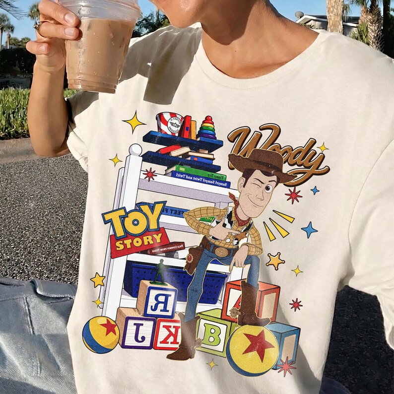 K&ouml;nnte beinhalten: Cremefarbenes T-Shirt mit einer farbenfrohen Grafik von Woody aus Toy Story. Das Design enth&auml;lt die W&ouml;rter "Toy Story" und "Woody" in einer verspielten Schriftart sowie Sterne und Spielzeugbl&ouml;cke. Das Shirt ist ein Freizeitkleidungsst&uuml;ck.