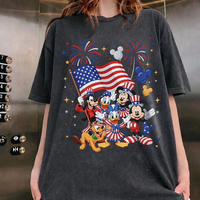 May include: Dark gray t-shirt featuring a patriotic design with Disney characters. The graphic includes Mickey Mouse, Minnie Mouse, Donald Duck, Goofy, and Pluto, along with an American flag and fireworks.