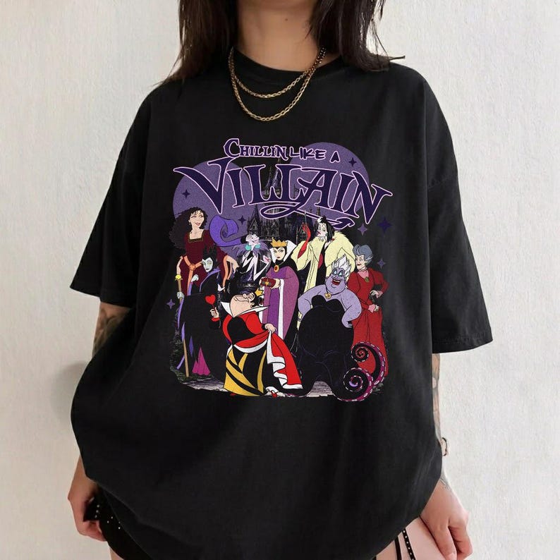 K&ouml;nnte beinhalten: Ein schwarzes kurz&auml;rmeliges T-Shirt mit einem Grafikdruck verschiedener animierter B&ouml;sewichte. Der Text "CHILLIN LIKE A VILLAIN" ist in Lila &uuml;ber den Charakteren aufgedruckt. Das Design zeigt mehrere Figuren in farbenfrohen Outfits.