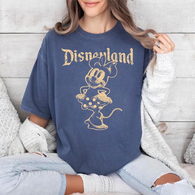 K&ouml;nnte beinhalten: Marineblaues T-Shirt mit einem Vintage-Grafik-Design von Minnie Mouse und dem Wort "Disneyland" in verblichener Goldfarbe. Das Design zeigt Minnie Mouse in einem gepunkteten Kleid. Das Shirt hat kurze &Auml;rmel und eine lockere Passform.