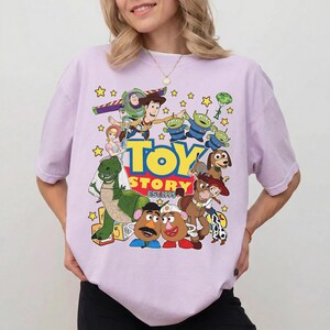 Pu&ograve; includere: Una maglietta lilla con una grafica vivace dei personaggi del film d'animazione "Toy Story". Il design include Buzz Lightyear, Woody e i piccoli omini verdi, con le parole "TOY STORY EST. 1995" in giallo e rosso.