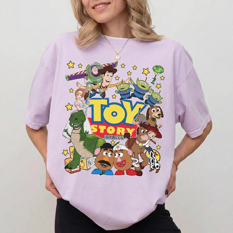 Pu&ograve; includere: Una maglietta lilla con una grafica vivace dei personaggi del film d'animazione "Toy Story". Il design include Buzz Lightyear, Woody e i piccoli omini verdi, con le parole "TOY STORY EST. 1995" in giallo e rosso.