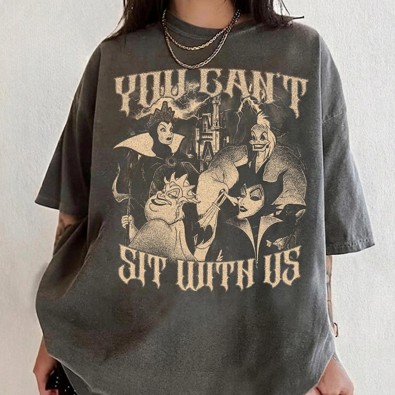 May include: Dark gray t-shirt featuring a graphic of Disney villains with the text "YOU CAN'T SIT WITH US". The design includes characters like Ursula, Maleficent, and the Evil Queen. The shirt has a vintage, distressed look.