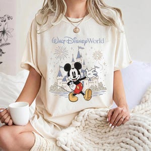 May include: Cream-colored t-shirt featuring a graphic of Mickey Mouse in front of a castle with fireworks and the text "Walt Disney World" in light blue. Mickey is wearing red shorts, yellow shoes, and black gloves.