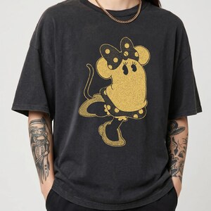 Pode incluir: T-shirt cinza escuro com um design dourado brilhante da Minnie Mouse. A personagem de desenho animado usa um la&ccedil;o e um vestido de bolinhas. A camisa tem mangas curtas e um ajuste folgado.