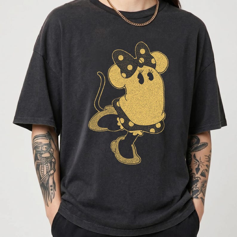 Pode incluir: T-shirt cinza escuro com um design dourado brilhante da Minnie Mouse. A personagem de desenho animado usa um la&ccedil;o e um vestido de bolinhas. A camisa tem mangas curtas e um ajuste folgado.