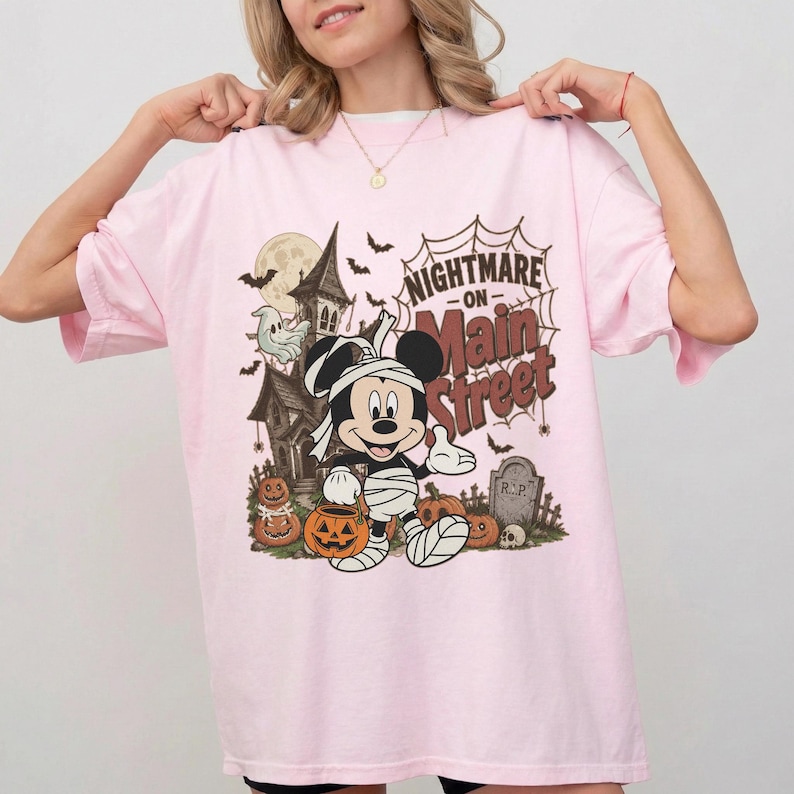 May include: Light pink t-shirt featuring a Halloween-themed graphic. The design includes a haunted house, bats, pumpkins, and a mummy-wrapped Mickey Mouse holding a jack-o'-lantern. The text "Nightmare on Main Street" is displayed above the character.