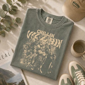 May include: A sage green short-sleeve t-shirt featuring a cream-colored graphic print of various animated villains with the text "CHILLIN LIKE A VILLAIN" above the characters. The shirt has a crew neckline and a Comfort Colors brand label inside.