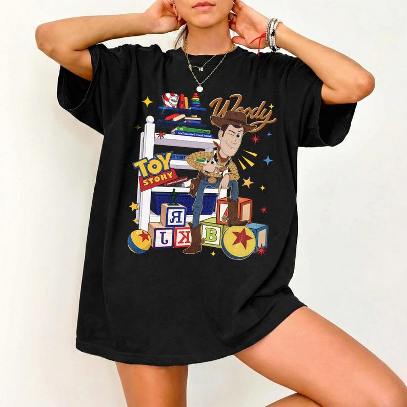 K&ouml;nnte beinhalten: Schwarzes T-Shirt mit einer Grafik von Woody aus Toy Story. Woody, mit Cowboyhut und Stiefeln, lehnt an einem Regal mit B&uuml;chern und Spielzeugbl&ouml;cken. Die Worte "Toy Story" und "Woody" sind ebenfalls abgebildet.