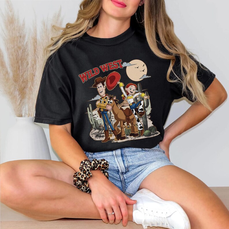 May include: Black t-shirt featuring a Wild West graphic with cartoon characters, a cowboy hat, and a full moon. The design includes Woody, Jessie, and Bullseye from Toy Story. The shirt is a casual, short-sleeved style.