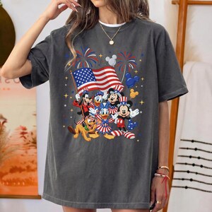 May include: Dark gray t-shirt featuring a patriotic design with Disney characters. The graphic includes Mickey Mouse, Donald Duck, Goofy, and Pluto, along with an American flag and fireworks. The shirt is a relaxed fit.