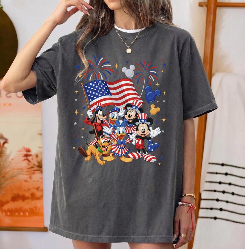 May include: Dark gray t-shirt featuring a patriotic design with Disney characters. The graphic includes Mickey Mouse, Donald Duck, Goofy, and Pluto, along with an American flag and fireworks. The shirt is a relaxed fit.