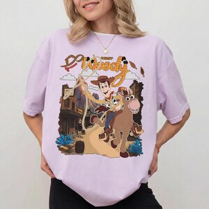May include: A light purple t-shirt featuring a cartoon graphic of Sheriff Woody from Toy Story riding a horse. The graphic includes the words "Sheriff Woody" in a western-style font, with a town scene in the background.