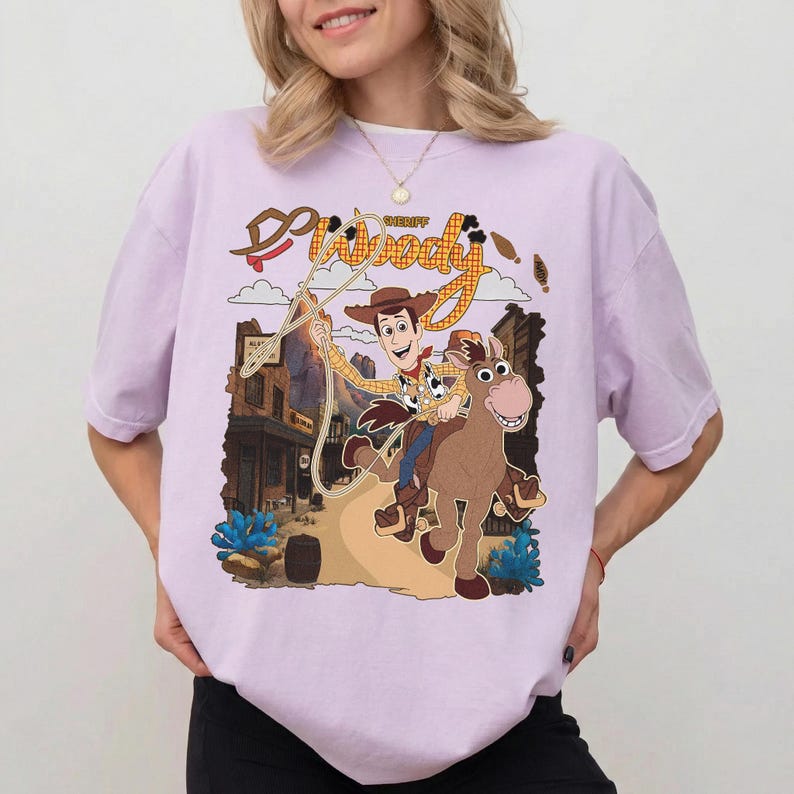 May include: A light purple t-shirt featuring a cartoon graphic of Sheriff Woody from Toy Story riding a horse. The graphic includes the words "Sheriff Woody" in a western-style font, with a town scene in the background.