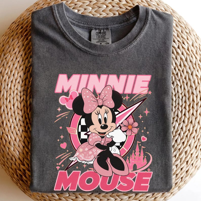 May include: Dark gray t-shirt featuring a graphic of Minnie Mouse in a pink dress and bow. The design includes the words "MINNIE MOUSE" in pink, a checkered flag, and a castle. The shirt is made of a soft material.