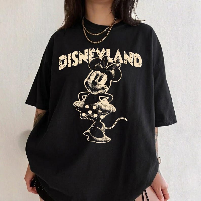 May include: Black t-shirt featuring a distressed graphic of Minnie Mouse and the word "Disneyland" in a vintage font. The design is in a cream color. The shirt has short sleeves and a relaxed fit.