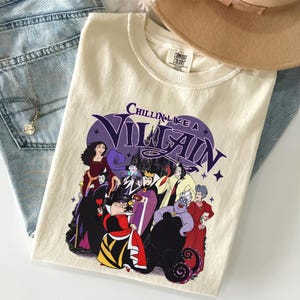 K&ouml;nnte beinhalten: Ein cremefarbenes Kurzarm-T-Shirt mit einem Grafikdruck verschiedener animierter B&ouml;sewichte. Der Text "CHILLIN LIKE A VILLAIN" ist in Lila &uuml;ber den Figuren aufgedruckt. Das T-Shirt hat ein Comfort Colors Markenetikett am inneren Halsausschnitt.