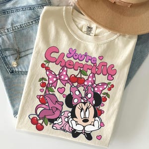 Pode incluir: Camiseta creme com um gr&aacute;fico da Minnie Mouse e o texto "You're Cherrific". O design inclui la&ccedil;os rosa, cerejas e cora&ccedil;&otilde;es. A camisa est&aacute; dobrada em cima de jeans azuis.