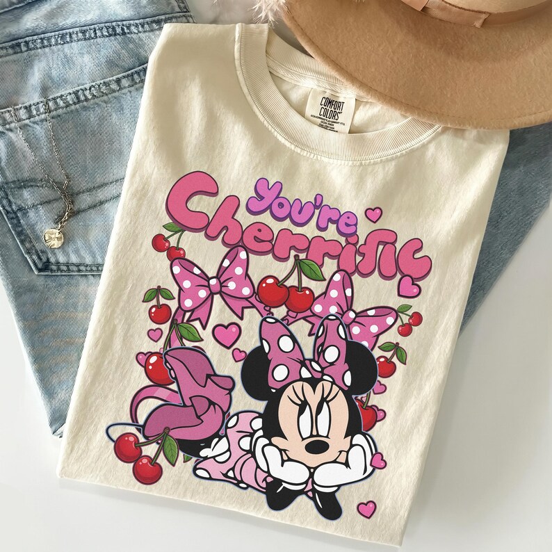 Pode incluir: Camiseta creme com um gr&aacute;fico da Minnie Mouse e o texto "You're Cherrific". O design inclui la&ccedil;os rosa, cerejas e cora&ccedil;&otilde;es. A camisa est&aacute; dobrada em cima de jeans azuis.