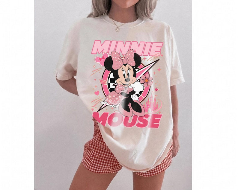 May include: Cream-colored t-shirt featuring a graphic of Minnie Mouse in a pink and black design. The text "MINNIE MOUSE" is displayed in pink. The shirt is paired with red and white checkered shorts.