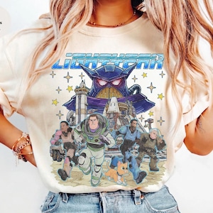 May include: Ivory-colored t-shirt featuring a colorful graphic of Buzz Lightyear and other characters from the movie Lightyear. The graphic includes the word "Lightyear" in blue block letters and a purple villain.