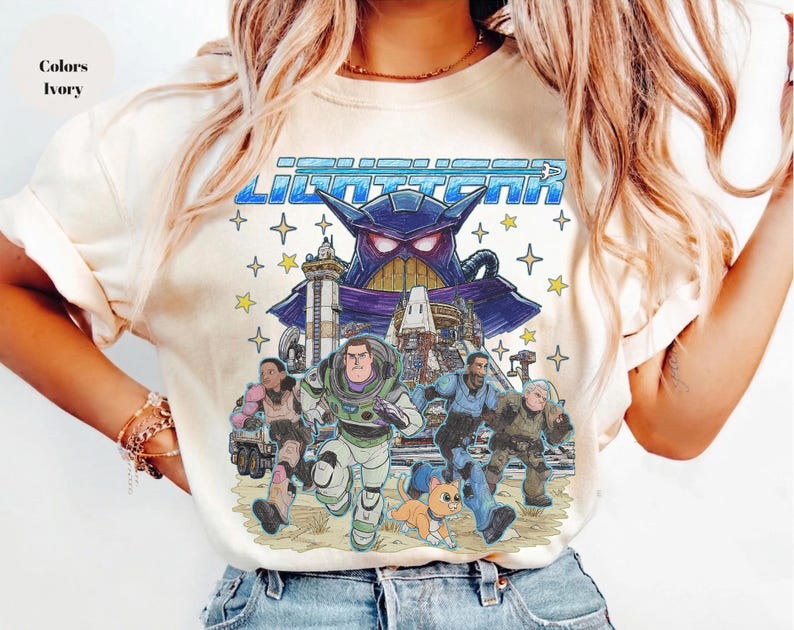 May include: Ivory-colored t-shirt featuring a colorful graphic of Buzz Lightyear and other characters from the movie Lightyear. The graphic includes the word "Lightyear" in blue block letters and a purple villain.