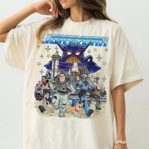May include: Cream-colored t-shirt featuring a colorful graphic of characters from the movie Lightyear. The design includes Buzz Lightyear, Zurg, and other characters, with the word "Lightyear" in blue letters across the top. The shirt has short sleeves.