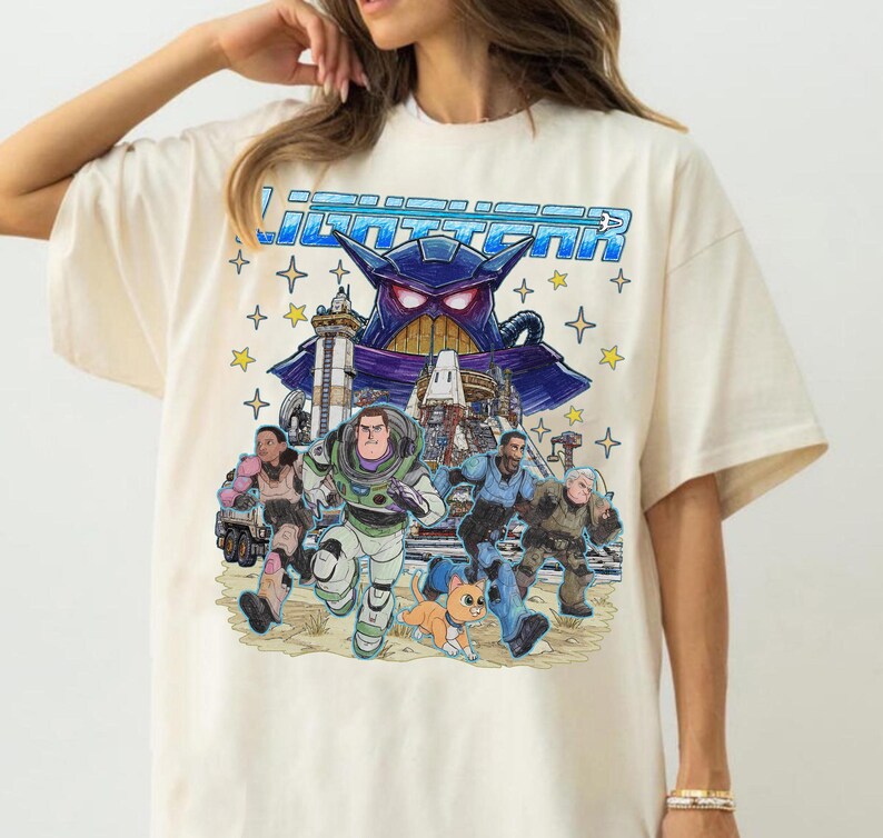 May include: Cream-colored t-shirt featuring a colorful graphic of characters from the movie Lightyear. The design includes Buzz Lightyear, Zurg, and other characters, with the word "Lightyear" in blue letters across the top. The shirt has short sleeves.