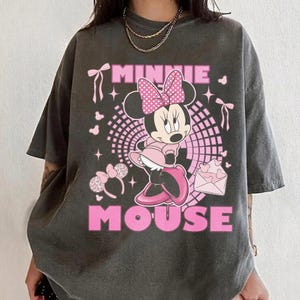 May include: Dark grey t-shirt with a pink Minnie Mouse design. The graphic includes Minnie in a pink dress and bow, with the words "MINNIE MOUSE" in pink. The shirt has a faded, vintage appearance.
