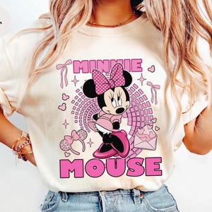 May include: Ivory t-shirt with a pink Minnie Mouse graphic. The design includes the words "MINNIE MOUSE" in pink, hearts, bows, and a polka-dot bow. The shirt is a casual style and is likely made of cotton.