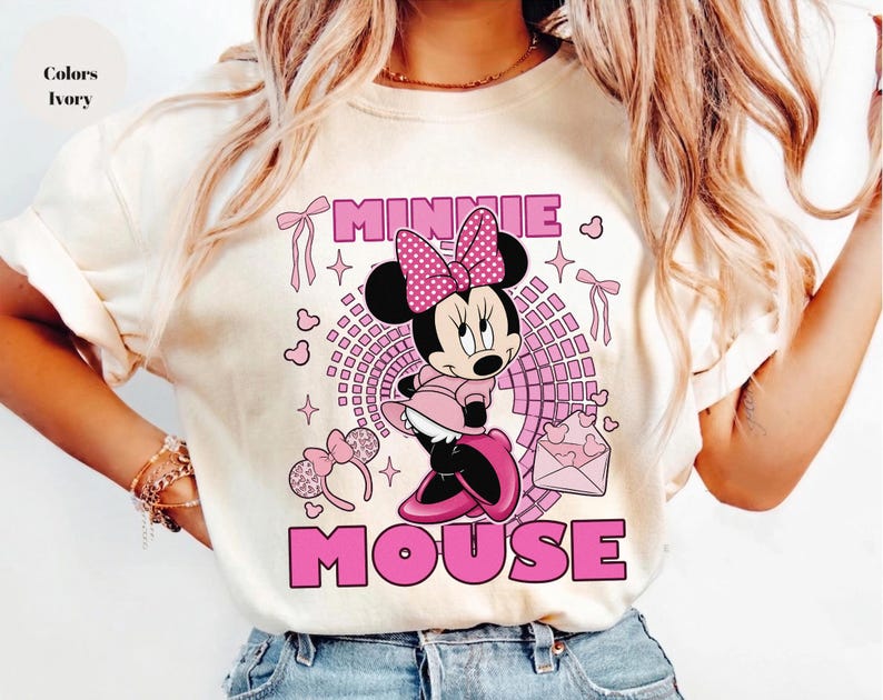 May include: Ivory t-shirt with a pink Minnie Mouse graphic. The design includes the words "MINNIE MOUSE" in pink, hearts, bows, and a polka-dot bow. The shirt is a casual style and is likely made of cotton.