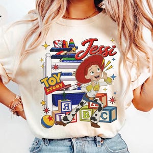 Puede incluir: Camiseta de color marfil con un dise&ntilde;o de Jessie de Toy Story. El dise&ntilde;o incluye la palabra "Jessie" en escritura roja, el logotipo de Toy Story y elementos tem&aacute;ticos de juguetes. La camiseta tiene cuello redondo.
