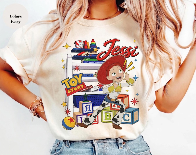 Puede incluir: Camiseta de color marfil con un dise&ntilde;o de Jessie de Toy Story. El dise&ntilde;o incluye la palabra "Jessie" en escritura roja, el logotipo de Toy Story y elementos tem&aacute;ticos de juguetes. La camiseta tiene cuello redondo.